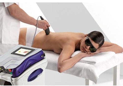 A male practitioner using a hand held medical laser to treat a female patient laying face down on a padded table